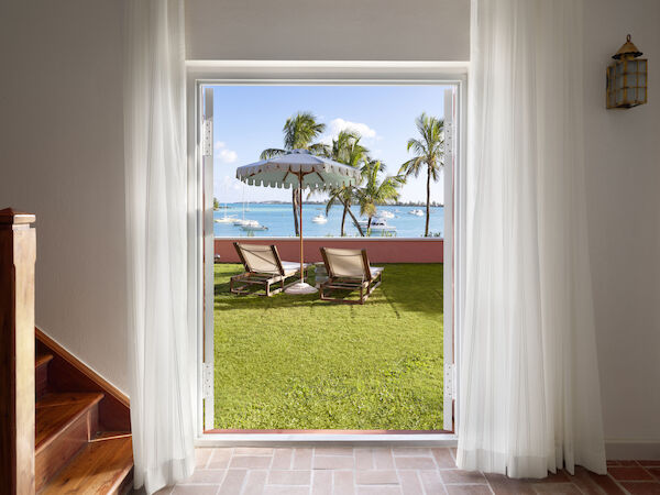A doorway opens to a view of a grassy yard with lounge chairs under an umbrella, overlooking a beach with palm trees and boats on the water.