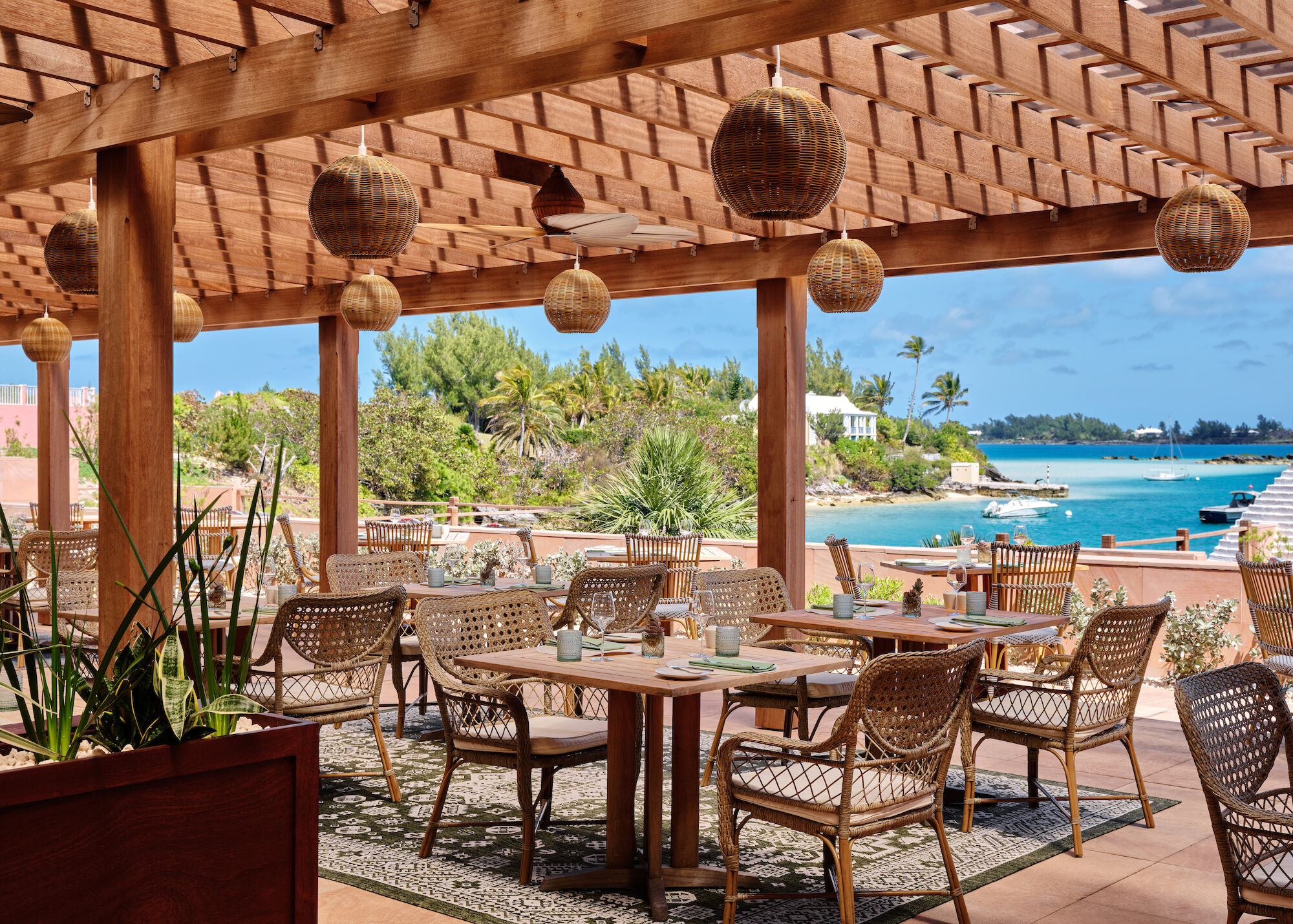 A seaside outdoor dining area with wicker chairs, round tables, and hanging lanterns under a wooden pergola, overlooking a blue bay.