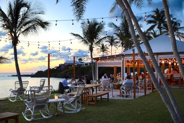 An outdoor dining area by the ocean at sunset, with palm trees, string lights, people dining, and a relaxing atmosphere.