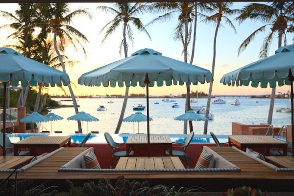 The image shows a scenic view of an outdoor seating area with umbrellas by the water, palm trees, and boats in the background at sunset.