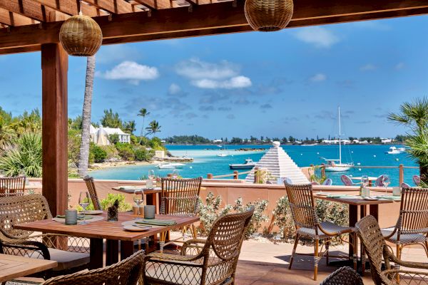 An outdoor dining area with wooden tables and wicker chairs overlooks a scenic beach with blue water, sailboats, and a clear sky.
