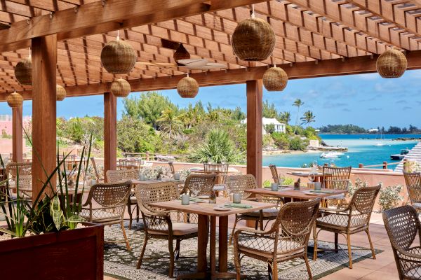 An outdoor restaurant or cafe with wicker chairs and wooden tables under a pergola, overlooking a serene waterfront with lush greenery.