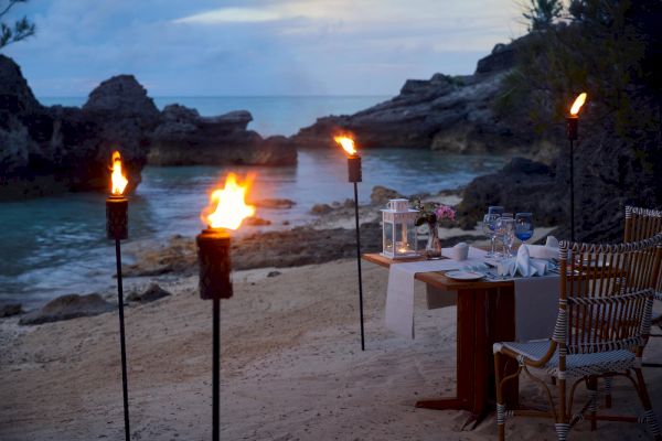 A romantic beachside dinner setup with torches, a table for two, and soft-lit lanterns, set against a backdrop of rocky shore and serene water.