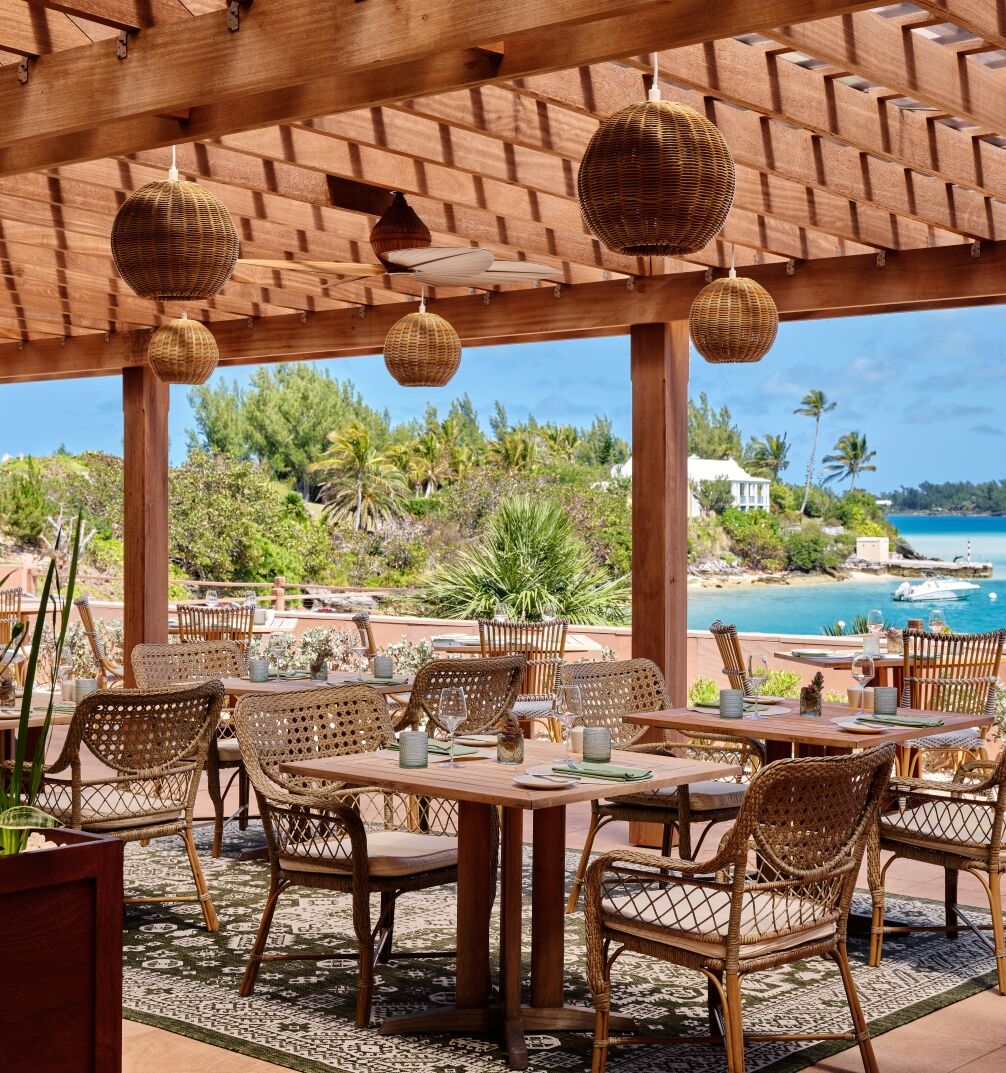 An outdoor seaside dining area with wicker chairs around tables under a wooden pergola, overlooking a blue ocean and small boats.