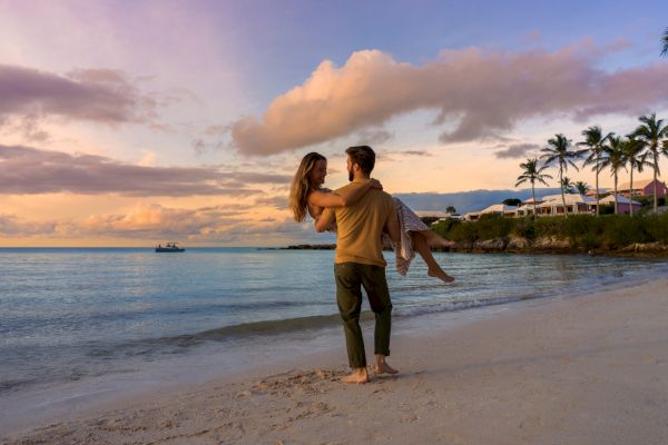 A couple is on a beach at sunset, with the man carrying the woman in his arms, with scenic ocean views and palm trees in the background.