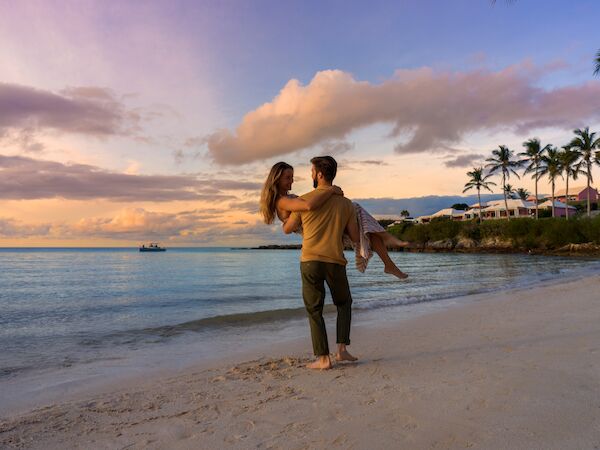 A couple is on a beach at sunset, with the man carrying the woman in his arms, with scenic ocean views and palm trees in the background.