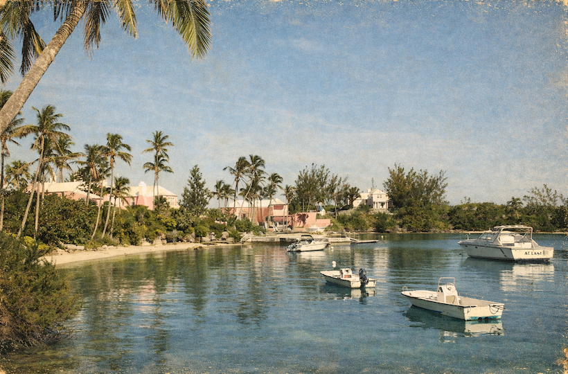 A tropical waterfront with palm trees, calm blue water, small boats near a sandy shore and pastel houses in the background, sunny and serene.