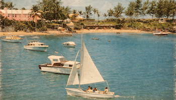 A sailboat glides through blue water near a beach with palm trees and several small boats; people relax aboard as a sunny coastal scene unfolds.