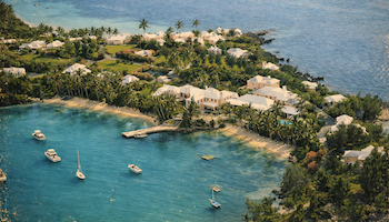 Tropical coastal resort with white villas among palm trees, a curved sandy bay, turquoise water, and small boats anchored near a rocky shoreline.