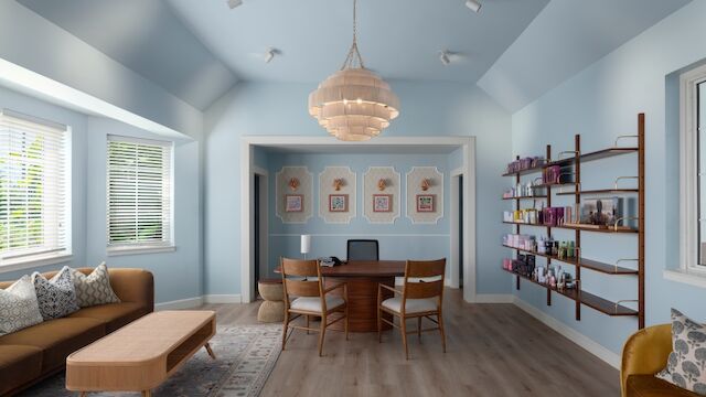 Bright, airy living area with light blue walls, a wooden dining table, shelving along the right, and a cozy sofa setup under a ceiling light.