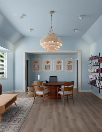 A bright, airy living room with light blue walls, a central chandelier, a wooden dining table, shelves with decor, and a cozy seating area with a sofa and armchairs.