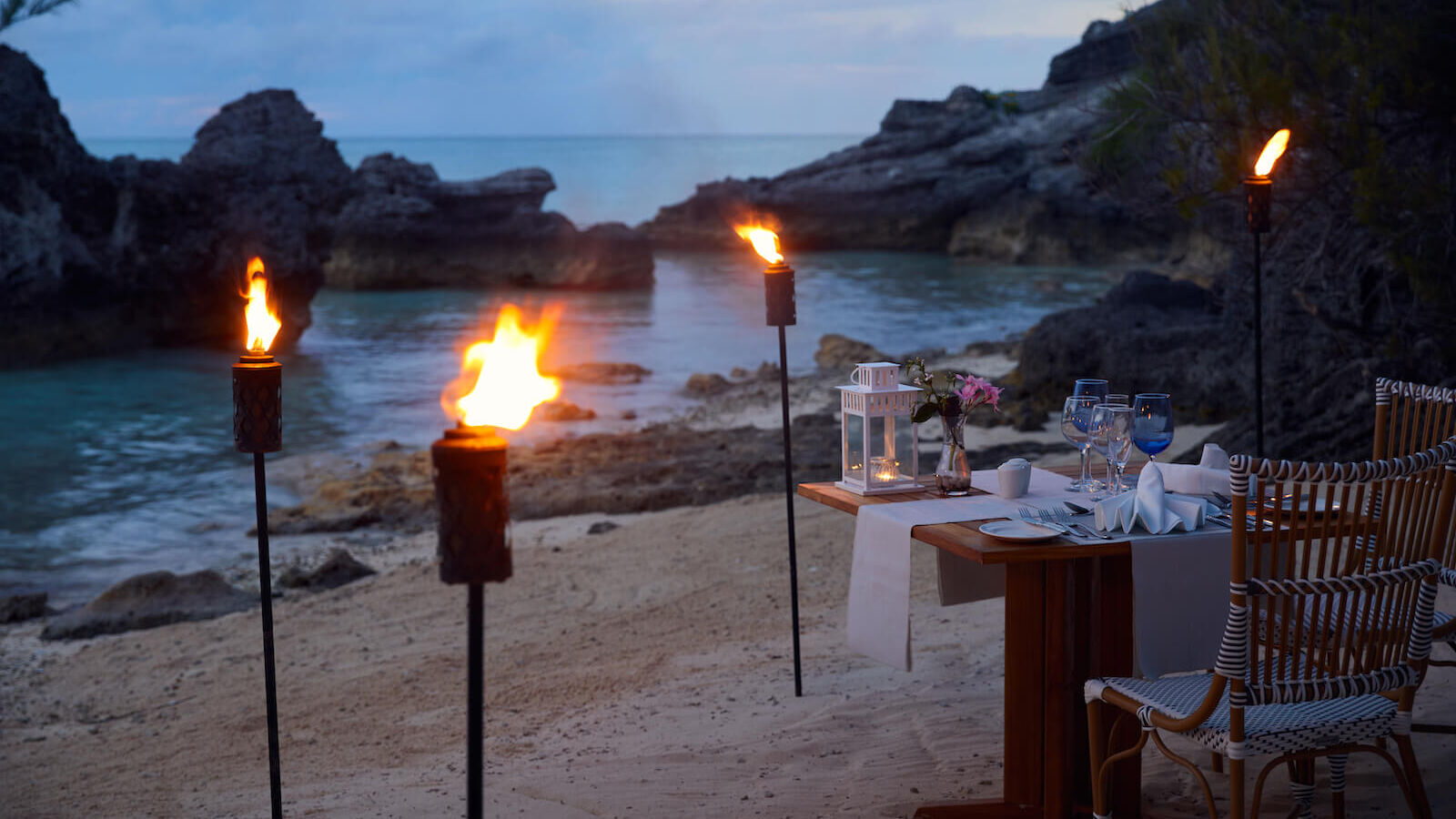 A romantic beachside dinner setup with torches, a table for two, and a view of the ocean surrounded by rocky cliffs at dusk.