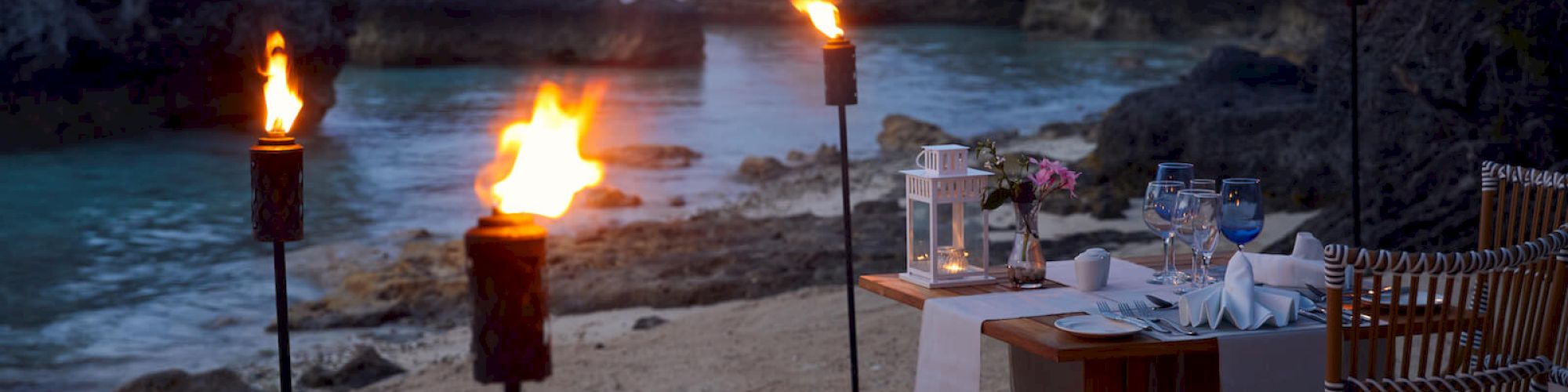 A romantic beachside dinner setup with torches, a table for two, and a view of the ocean surrounded by rocky cliffs at dusk.