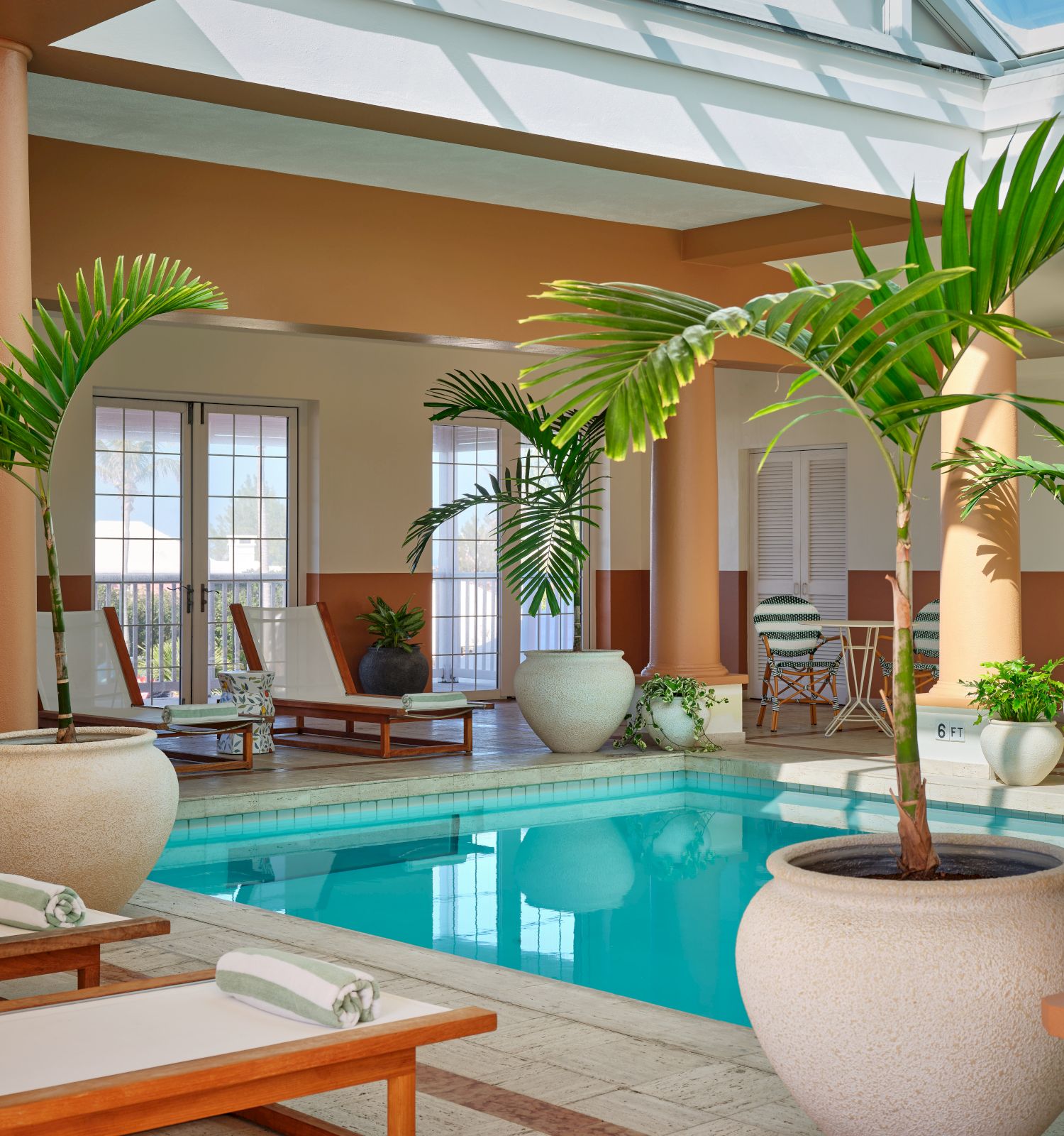 The image shows an indoor pool area with potted palm trees, lounge chairs, and large windows letting in natural light.