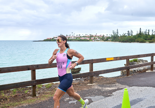 A female triathlete runs along a coastal seawall in a pink and blue racing suit, wearing sunglasses, with a wooden fence, turquoise water, and a distant shoreline in the background.