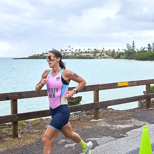 A female triathlete runs along a coastal seawall in a pink and blue racing suit, wearing sunglasses, with a wooden fence, turquoise water, and a distant shoreline in the background.