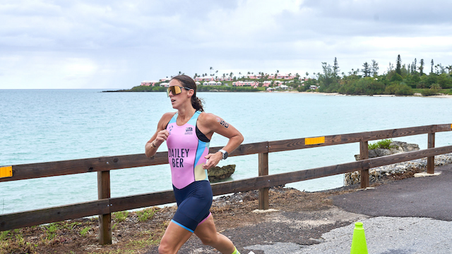 A female triathlete runs along a coastal seawall in a pink and blue racing suit, wearing sunglasses, with a wooden fence, turquoise water, and a distant shoreline in the background.