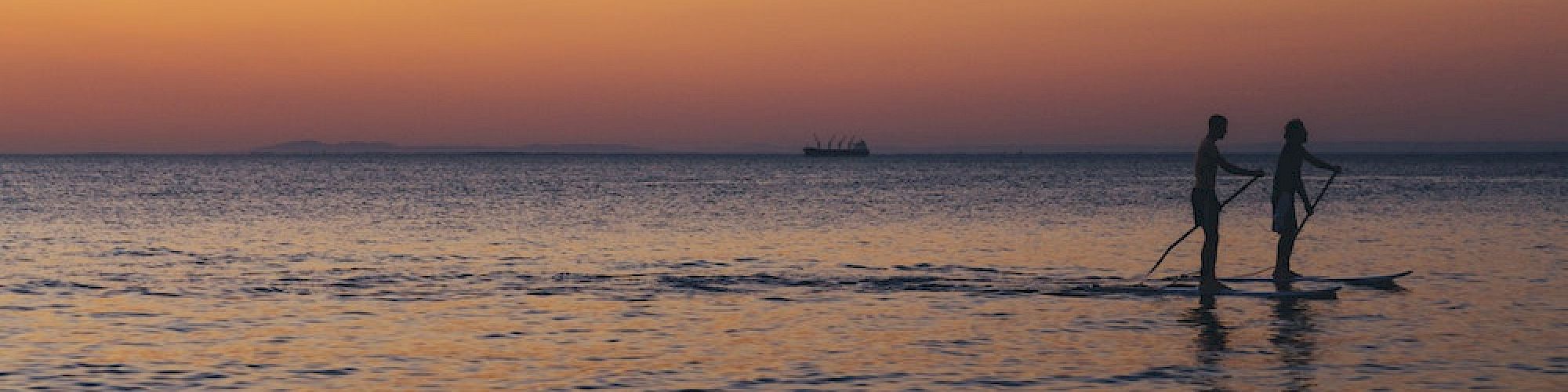 Two people are paddleboarding on calm water during a serene sunset, with a distant ship silhouette on the horizon.