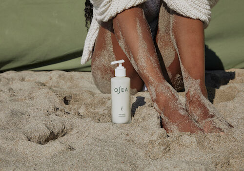 A bottle of OEA lotion on sandy beach next to a person&rsquo;s sandy legs, feet resting in the sand, with a towel or wrap nearby.