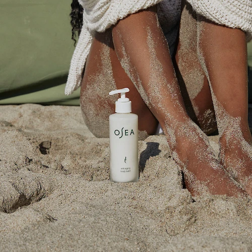 A bottle of OEA lotion on sandy beach next to a person’s sandy legs, feet resting in the sand, with a towel or wrap nearby.