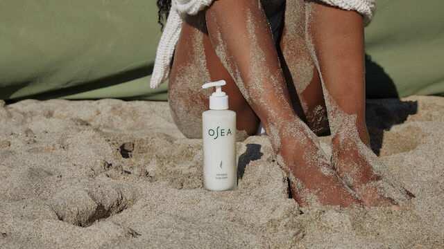 A bottle of OEA lotion on sandy beach next to a person&rsquo;s sandy legs, feet resting in the sand, with a towel or wrap nearby.
