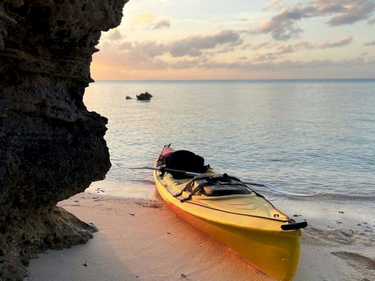 A yellow kayak is resting on a sandy beach near a rocky outcrop, with a calm sea and a setting sun in the background.