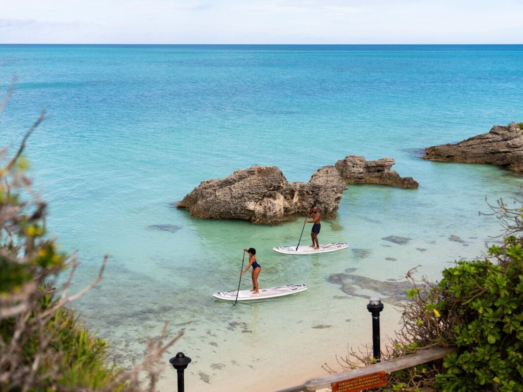 Two people stand on paddleboards near a rocky shoreline as turquoise water meets a sandy beach at a sunny coastal scene.
