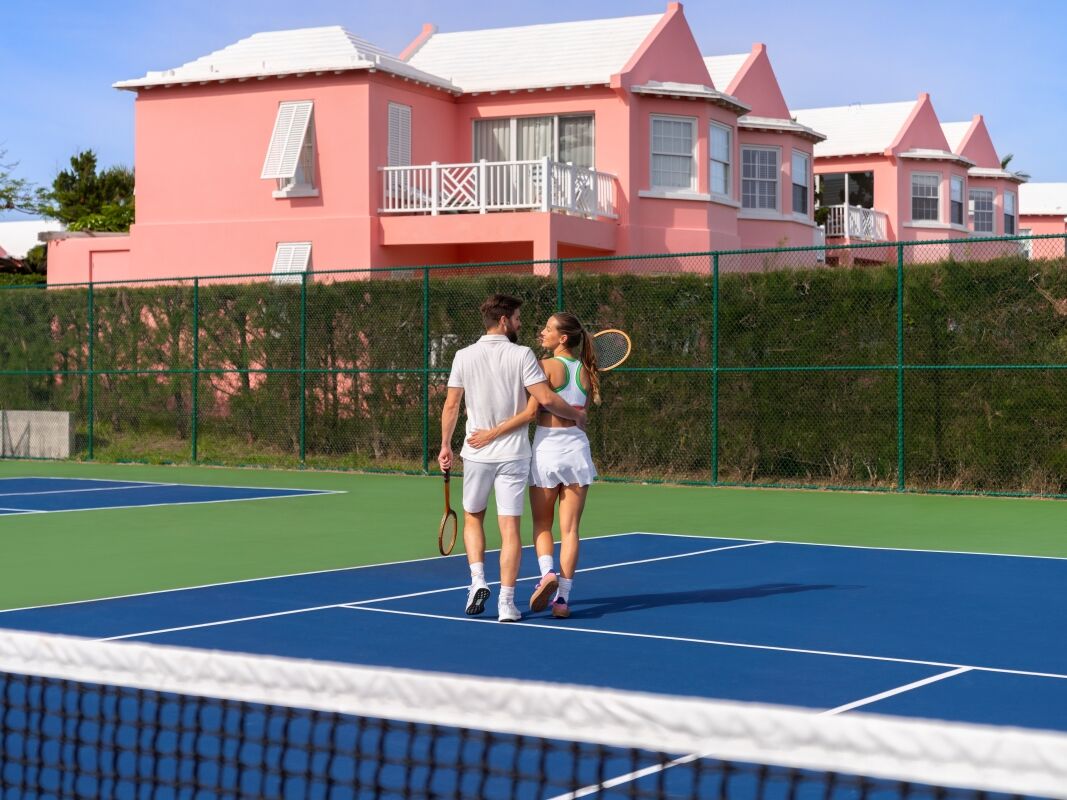 A couple walks hand-in-hand on a blue tennis court near pink buildings, with a net in the foreground and a bright, sunny sky.