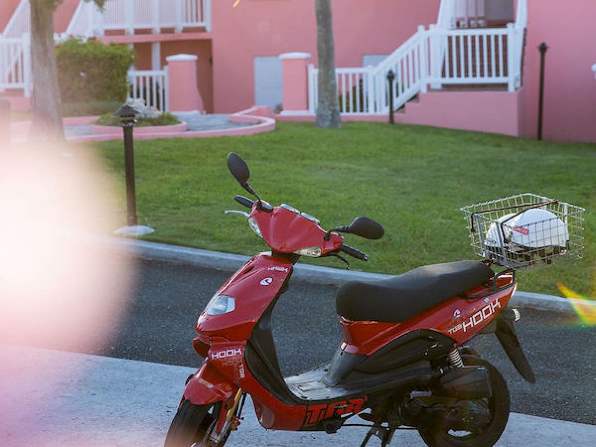 A red scooter is parked in front of pink buildings, green lawn, palm trees, and with a mountain backdrop. Sunlight flares in the top left corner.