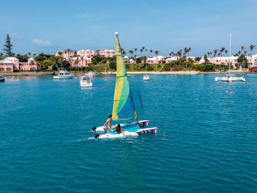 A small sailboat with a bright yellow-green sail glides on calm turquoise water near a coastal town with pastel buildings and palm trees.