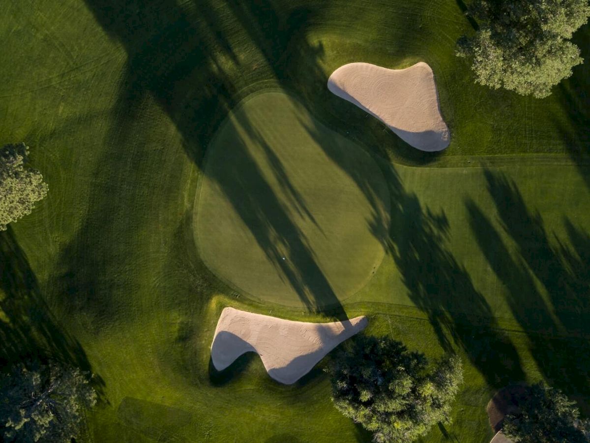 An aerial view of a golf course showing two sand traps adjacent to a green, surrounded by trees casting long shadows.