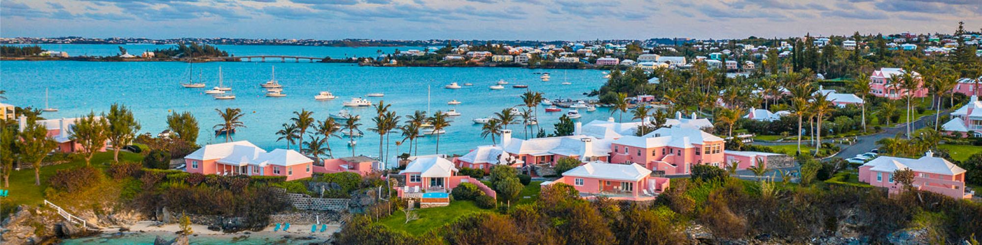 A coastal scene with turquoise waters, pink houses, palm trees, and sailboats anchored in a calm bay on a sunny day paired with a cloudy sky.