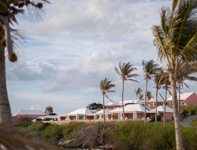 A coastal scene with palm trees, a sandy beach, and red-roofed buildings in the background under a partly cloudy sky.