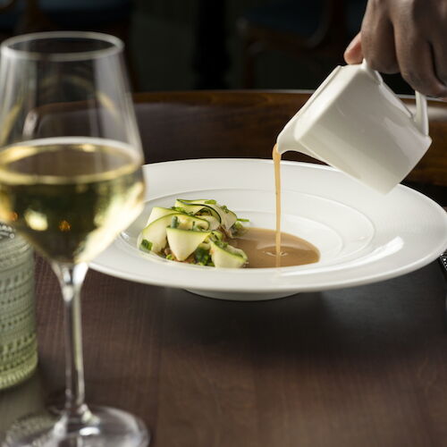 A chef pours warm sauce onto a plated salad in a white bowl at a dining table, with wine glasses nearby.