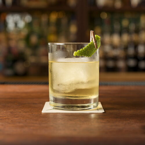 A pint of pale beer sits on a bar counter, topped with a green garnish and a slice of lime, in a cozy pub setting.