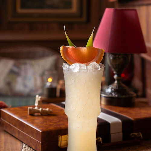 A tall iced cocktail topped with an orange-melon garnish and green leaves, served on a bar table with a pink lamp in back.