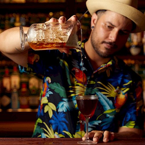 A bartender in a tropical shirt and a hat is pouring a drink from a jug into a glass at a bar with shelves of bottles in the background.