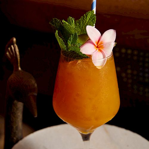 An orange cocktail garnished with mint leaves, a pink flower, and a striped straw, served in a stemmed glass on a round white surface.