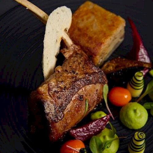 A gourmet dish featuring a lamb chop, a pastry square, green sauce, cherry tomatoes, and artistic vegetable garnishes on a black plate.