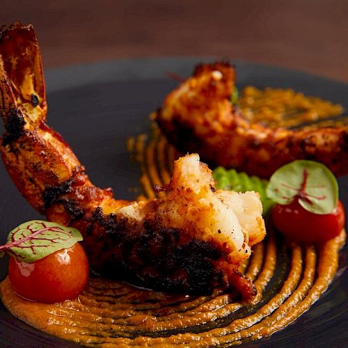 The image shows a plate with grilled shrimp, cherry tomatoes, green leaf garnish, and a swirl of sauce, likely a gourmet dish.