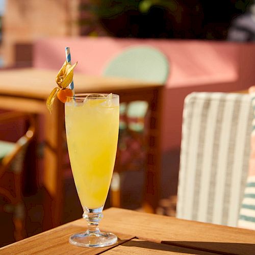 A tall glass of yellow drink with a straw and garnish on a wooden table, with a blurred outdoor patio setting in the background, in daylight.