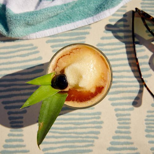 A tropical cocktail garnished with green leaves and a cherry sits on a patterned surface, next to a pair of sunglasses and a towel.