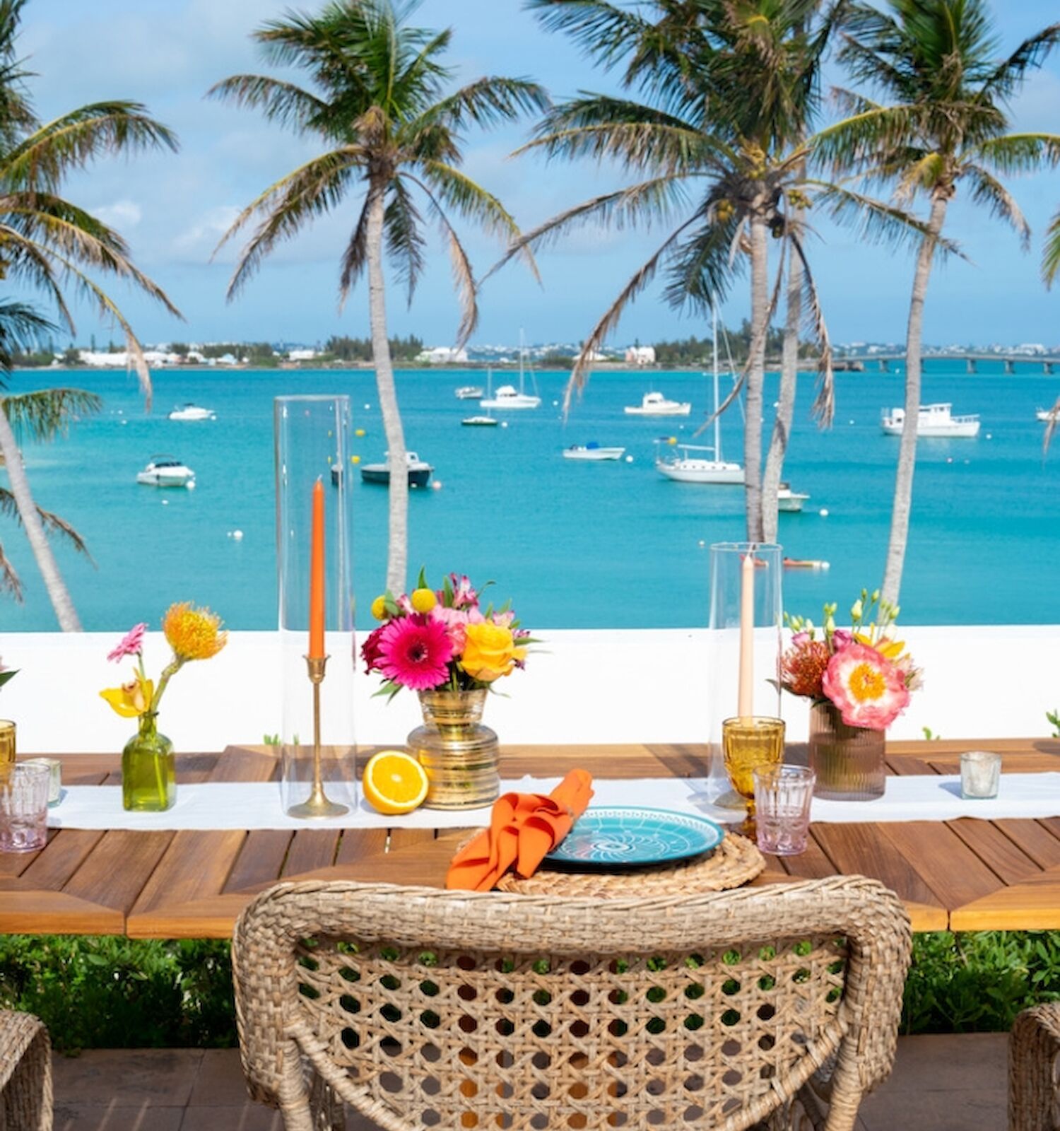A decorated outdoor table with chairs is set up for dining, overlooking a turquoise ocean with boats and tall palm trees under a clear blue sky.