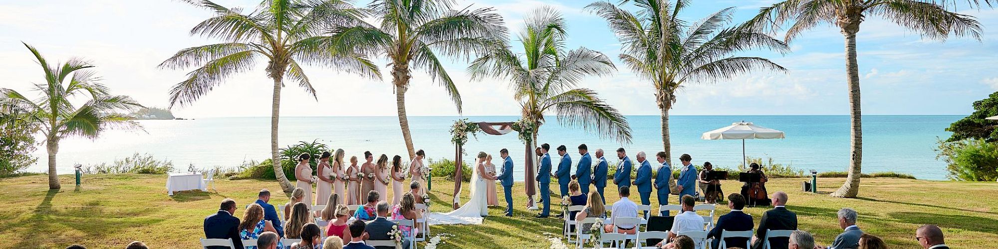 Guests seated at an outdoor wedding ceremony by the ocean, lined with palm trees, and a couple standing at the altar with the bridal party.