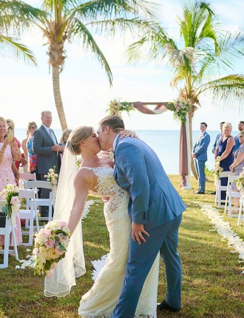 A newlywed couple shares a kiss under palm trees at an outdoor wedding ceremony surrounded by guests in a picturesque setting.