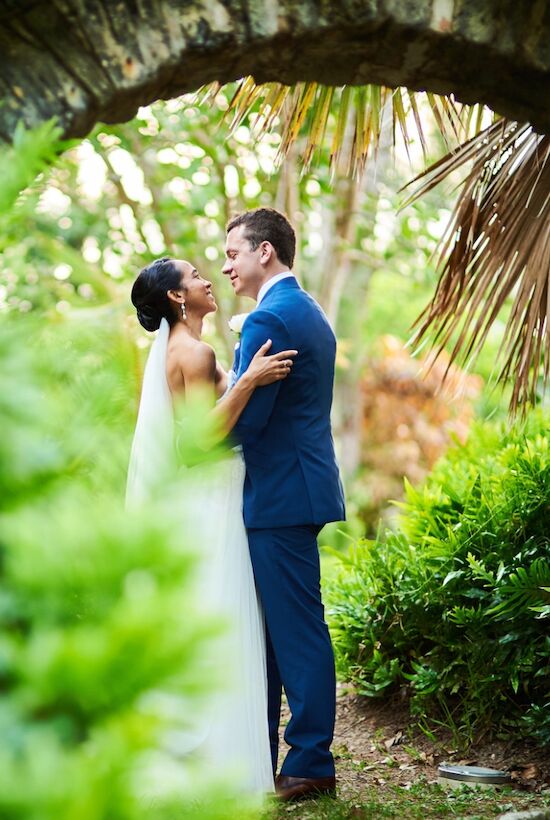 A bride and groom embrace lovingly in a lush, green garden setting, beneath a stone archway, sharing a special moment on their wedding day.
