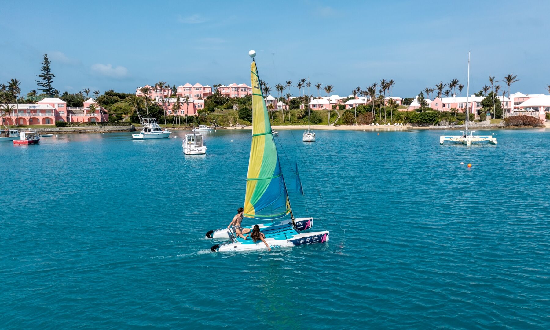 Sailboat with a bright yellow-green sail rides clear blue water near a harbor, pastel buildings, palm trees, and small boats in the background.