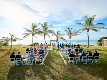 An outdoor wedding ceremony is taking place under palm trees near the ocean, with guests seated in rows and the bridal party at the altar.