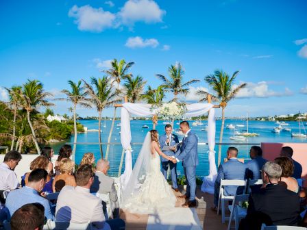 A wedding ceremony by a waterfront; a couple stands under an arch adorned with flowers, surrounded by seated guests. Palms and boats in the background.
