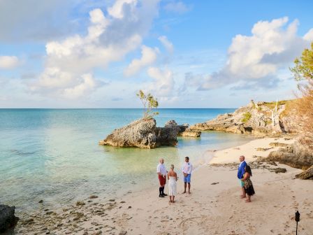 A group of five people stands on a picturesque beach with clear blue water and rocks, under a partly cloudy sky.